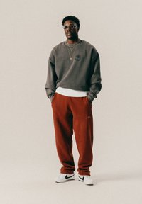 Άνδρας στέκεται με τα χέρια στις τσέπες, φοράει γυαλιά, γκρι φούτερ, κόκκινες παντελόνες, λευκά αθλητικά παπούτσια Nike και στρωτές χρυσές αλυσίδες σε απλό φόντο.