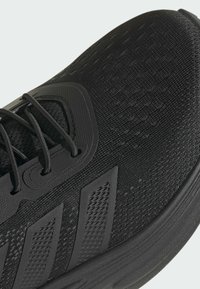 Close-up of black athletic shoe with textured mesh fabric, lace-up design, and three patterned stripes on the side.