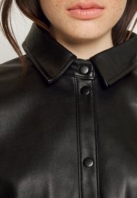 Svart läderjacka med en stor krage, försedda med svarta tryckknappar och en texturerad yta. Nära bild av tyget och detaljhanden.