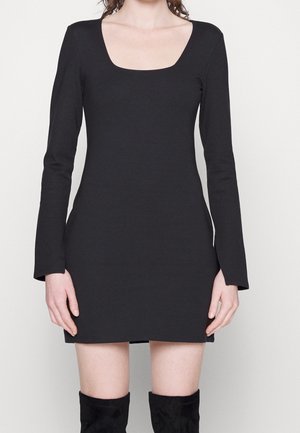 Kvinde iført en sort, langærmet, tætsiddende minikjole med firkantet udskæring og sorte knælange støvler mod en neutral baggrund.