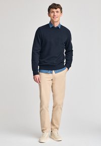 Felpa blu scuro con logo "POLO CLUB" in rilievo, sovrapposta a una camicia di denim azzurra. Abbinata a pantaloni beige e sneaker color crema.