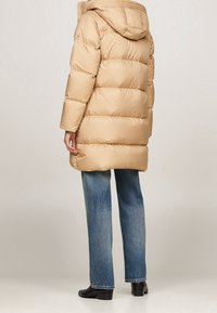 Beige pufferjack met een capuchon, voorzien van een gestikte textuur en een losse pasvorm, gecombineerd met blauwe rechte jeans en zwarte enkellaarsjes.