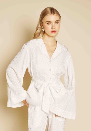 Blondýna v bílé saténové pyžamové soupravě s puntíky, s páskem v pase a náušnicemi ve tvaru srdce, stojící na béžovém pozadí.