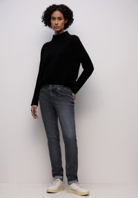 Pull noir à col haut associé à un jean gris décoloré slim et des baskets blanches avec des accents crème. Fond uni.