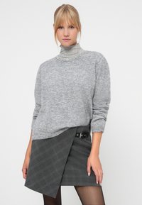 Grå strikket sweater med høj hals og dekorativ krave. Kombineret med en grå ternet indpakningsnederdel med en sidelukning.