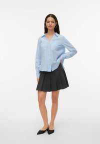 Chemise bleu clair à boutons avec col, dotée de deux poches poitrine, associée à une jupe plissée gris foncé et des ballerines noires pointues.