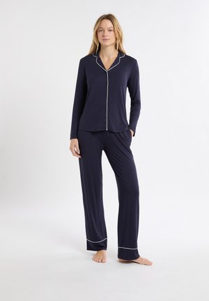 Femme debout pieds nus portant un haut de pyjama bleu marine à manches longues et boutons, avec un pantalon assorti comportant une bordure blanche sur le col et les poignets.