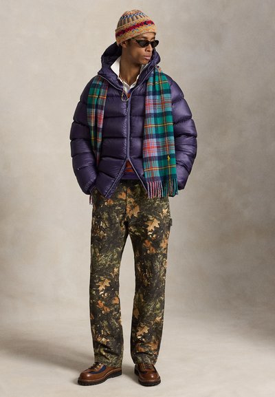 Man wearing purple puffer jacket, colorful knit hat, plaid scarf, camouflage pants, brown shoes, and black sunglasses standing against neutral background.