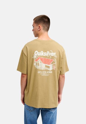 Homme portant un t-shirt beige Quiksilver avec un motif de maison de plage au toit rouge et un texte au dos, associé à un jean bleu, debout de dos.