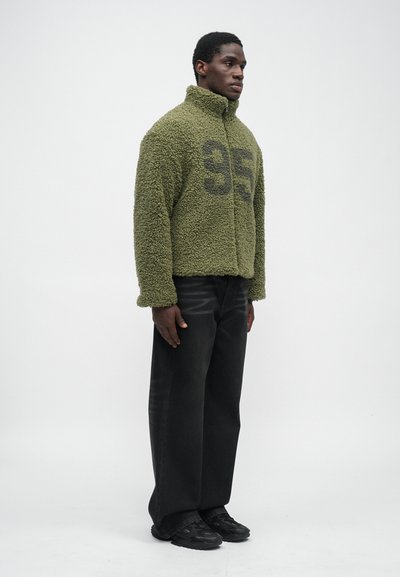 Green fleece jacket with a zip closure, featuring a large black '95' print on the front. Paired with loose black pants and black footwear.
