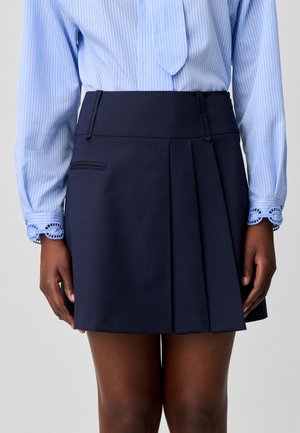 Mujer con falda plisada azul marino y blusa de rayas azul claro con detalle de encaje en los puños, de pie frente a un fondo liso.