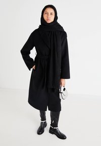 Svart, lang frakk med belte, parret med et tykt svart skjerf. Svart knelang støvler og en stripete håndveske fullfører antrekket.