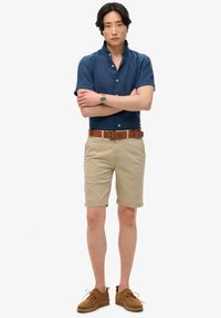 Hombre de pie con los brazos cruzados, vestido con una camisa azul de manga corta, pantalones cortos beige, cinturón marrón, reloj marrón y zapatos de ante marrones, sobre un fondo blanco.