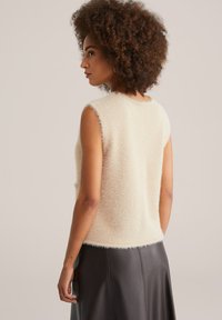 Top senza maniche in crema fluffy con vestibilità rilassata, caratterizzato da una texture morbida e orli grezzi, abbinato a una gonna in pelle scura.