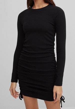 Femme portant une mini-robe noire cintrée à manches longues avec fronces et liens sur les côtés, debout devant un fond gris uni.