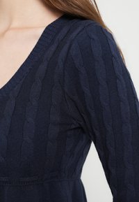 Μπλε ναυτικό μάλλινο πουλόβερ με V-λαιμόκοψη, με ανάγλυφο σχέδιο καλωδίου κατά μήκος των μανικιών και του σώματος, κατασκευασμένο από απαλό ύφασμα.