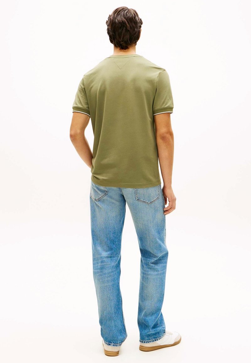 Homme debout, dos face à la caméra, portant une chemise à manches courtes olive, un jean bleu clair et des baskets blanches sur fond blanc.