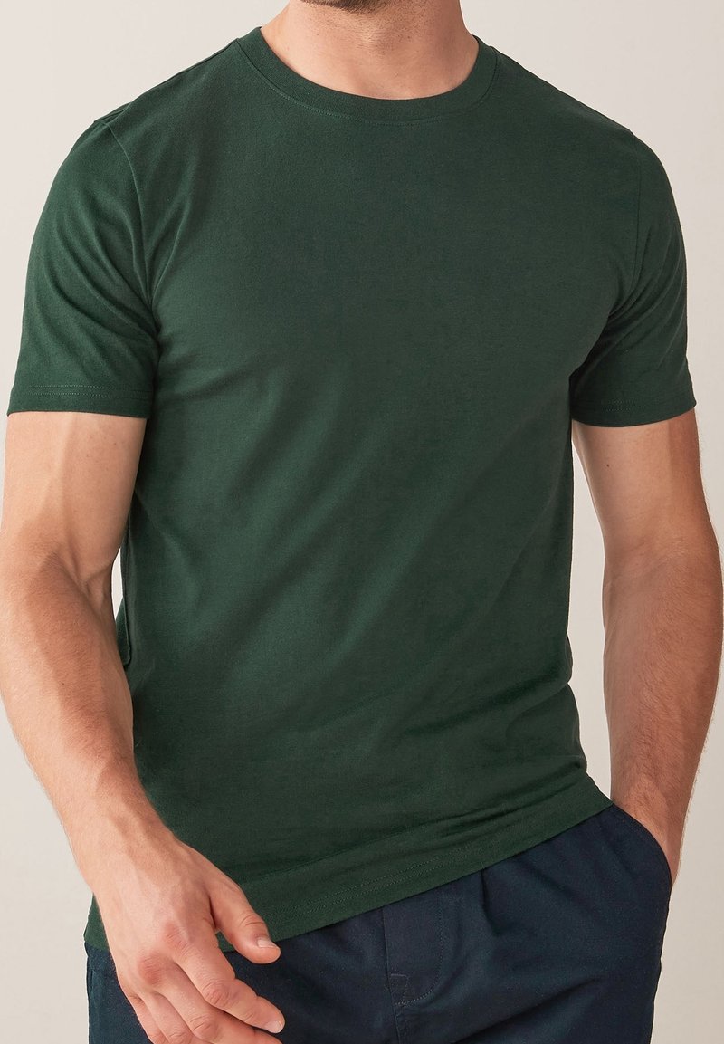 Homme portant un t-shirt uni vert foncé à manches courtes avec un col rond et un pantalon bleu foncé, debout avec une main dans la poche.