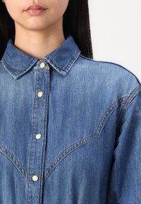 Denim srajca z ostrim ovratnikom, gumbom spredaj z bledejšimi gumbi in zahodnim stilom yoke detajli v modri tkanini.
