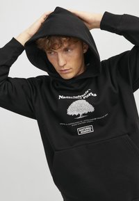 Schwarzer Kapuzenpullover aus Baumwolle. Mit einem Baum-Graphic und weißem Text, sowie einer Tasche auf der Vorderseite. Glatte Textur.
