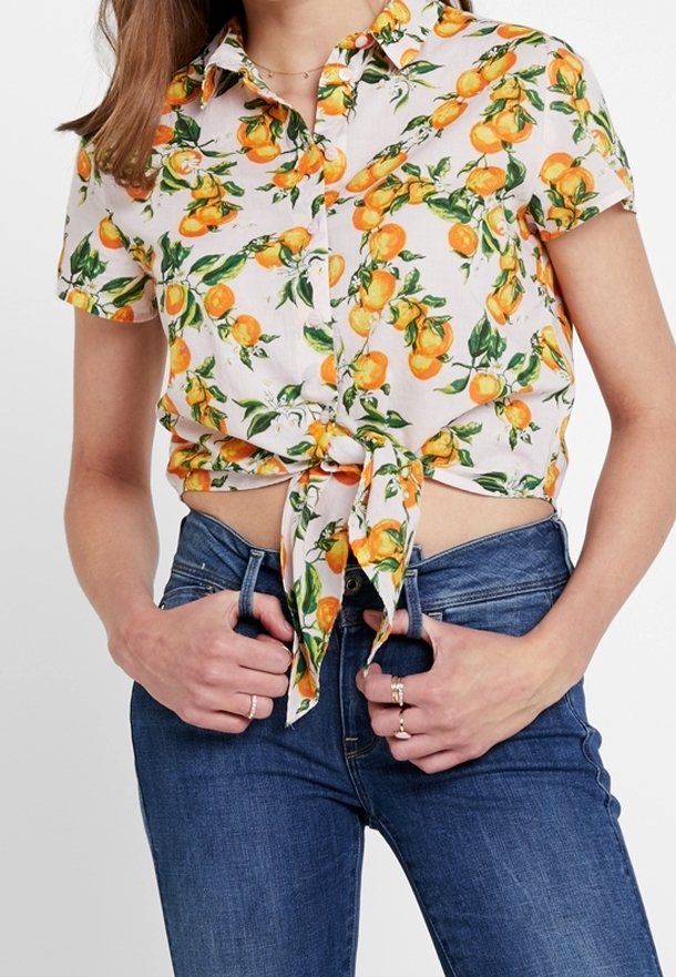Femme portant une chemise à manches courtes à boutons avec imprimé orange nouée à la taille et un jean bleu, mains dans les passants de ceinture.
