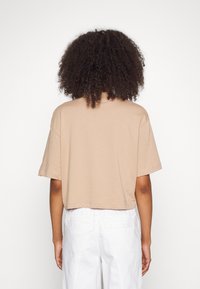 Osoba s kudrnatými vlasy, která má na sobě béžovou krátkou vyztuženou košili s krátkým rukávem a bílé kalhoty, stojí před jednobarevným bílým pozadím.