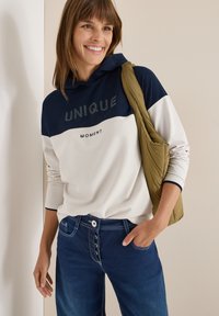 Hoodie color block bianco e navy con testo 'UNIQUE', abbinato a jeans scuri in denim con chiusura a bottoni e tasche. Borsa a spalla verde oliva.