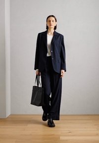 Abito a righe navy con giacca strutturata e pantaloni a gamba larga, abbinato a una maglietta bianca e una borsa tote nera. Tessuto liscio, design minimalista.