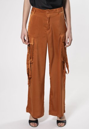 Pantaloni cargo in satin arancione con vita alta, due tasche laterali e dettagli legati ai lati, abbinati a scarpe nere con la punta aperta.