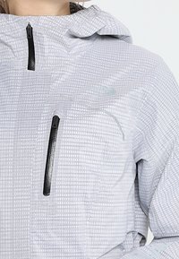 Jasnoszary wzorzysty kurtka z czarnymi zamkami błyskawicznymi z przodu i na klatce piersiowej oraz kapturem, noszona przez osobę.