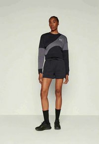 Žena oblečená v černé a šedé sportovní mikině, černých kraťasech, černých ponožkách a černých teniskách, stojící proti jednobarevnému světlému pozadí.