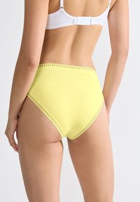 Vrouw in hoge gele onderbroek met kantafwerking en een witte beha, gezien vanachter tegen een effen achtergrond.