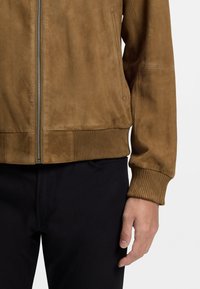 Brun mockabomberjacka med ribbade muddar och fåll, utrustad med dragkedja framtill och sidofickor. Kombinerad med mörka byxor.