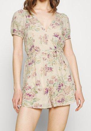 Femme portant une combinaison courte crème à fleurs avec manches bouffantes, col en V et ceinture à la taille, debout devant un fond uni.