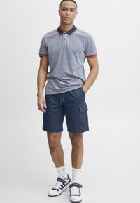 Tricou polo gri cu accente roșii și bleumarin, asortat cu pantaloni scurți cargo bleumarin. Sosete albe și teneși completează ținuta. Outift casual în general.