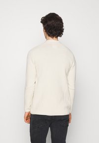 Mann mit dunklem, lockigem Haar, der ein cremefarbenes, geripptes Langarmshirt und schwarze Jeans trägt, steht vor einem schlichten weißen Hintergrund.