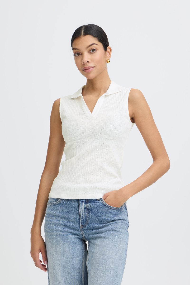 Femme aux cheveux foncés en chignon bas, portant des boucles d'oreilles créoles en or, un haut en maille blanc sans manches avec col, et un jean bleu clair avec une main dans la poche.