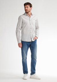 Uomo che indossa una camicia bianca a righe con bottoni, jeans blu e sneakers bianche, in piedi con una mano in tasca, mentre guarda di lato.