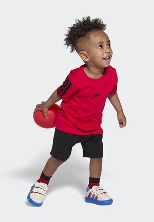 Rode katoenen T-shirt met zwart Adidas-logo en drie strepen, zwarte shorts en wit-blauwe sneakers, een rode bal vasthoudend, glimlachend.
