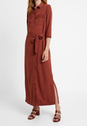 Vrouw draagt een lange, roestkleurige jurk met knopen, borstzakken, een ceintuur, een zijsplit en bruine sandalen met hak.