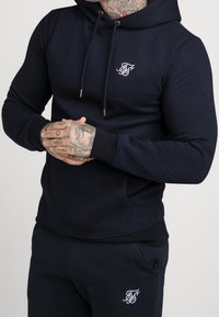 Uomo in felpa blu navy e pantaloni abbinati con logo ricamato bianco, che mostra le mani tatuate giunte davanti.