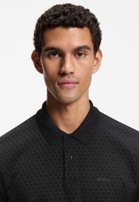 Schwarzes Poloshirt mit hexagonalem Muster, gefertigt aus strukturiertem Stoff. Verfügt über einen Kragen, zwei Knöpfe und das "BOSS"-Logo auf der Vorderseite.
