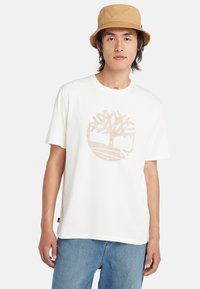 T-shirt en coton blanc avec un logo graphique d'arbre brun clair. Le design présente un col rond et des manches courtes, associé à un jean bleu.