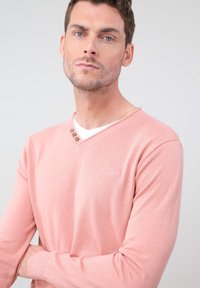 Pull rose à manches longues avec un col en V, arborant trois boutons décoratifs au niveau du col et un petit logo. Matière douce et coupe ajustée.