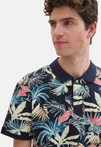 Kortærmet polo-shirt i marineblå med et tropisk bladmønster i blå, grøn og pink. Har en krave og tre knapper ved åbningen.