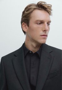 Blazer nero sopra una camicia scura, con revers a notched, taschino singolo e texture opaca, su uno sfondo neutro.