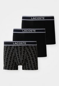 Trois paires de boxers noirs avec un logo Lacoste blanc sur la ceinture. La paire de devant présente un motif géométrique en gris.