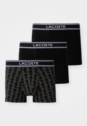Trzy pary czarnych bokserek z białym logo Lacoste na gumce. Przód jednej z par ozdobiony jest geometrycznym wzorem w odcieniach szarości.