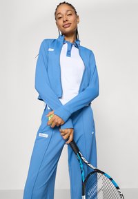 Lysblå atletisk jakke med lynlås og matchende bukser, med en hvid skjorte under. Indeholder en tennisracket og bold.