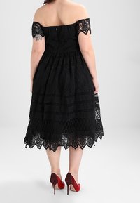 Černé šaty bez ramínek z krajky s těsně přiléhajícím topem, vrstvenou sukní, zvlněným lemem a podrobnými krajkovými vzory, doplněné červenými podpatky.